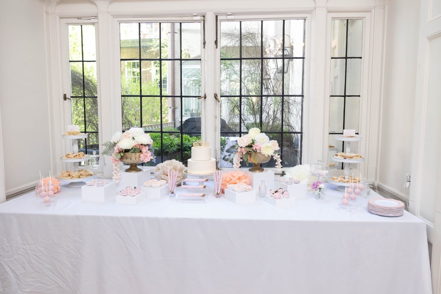 Dessert table against floor-to-ceiling windows \u2014 two-tier cake, tiered trays, cake pops, floral arrangements in gold urns