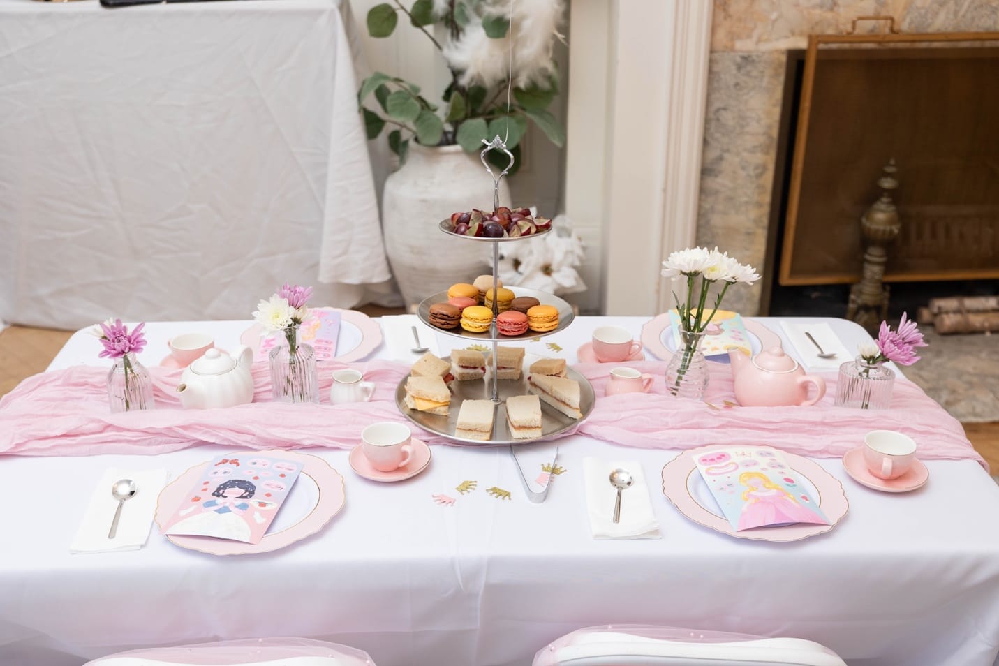 Close-up of a single table setting \u2014 porcelain teacups, tiered stand with macarons and finger sandwiches, bud vases with fresh flowers